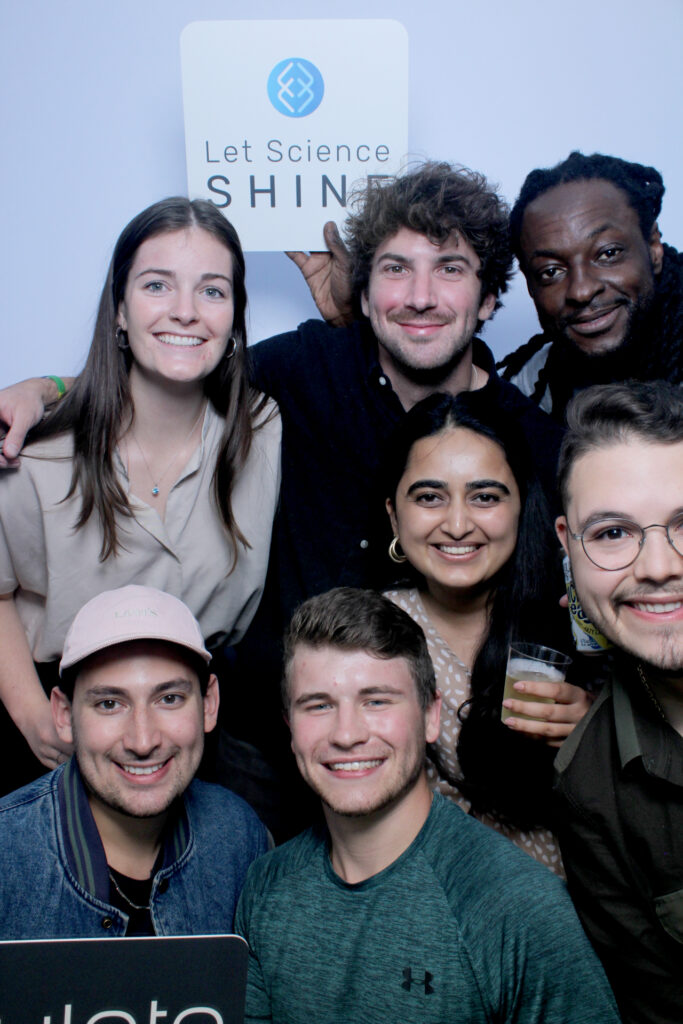 A group of people smiling into the camera. One is holding a sign printed with the Emulate logo and the words "Let Science Shine".